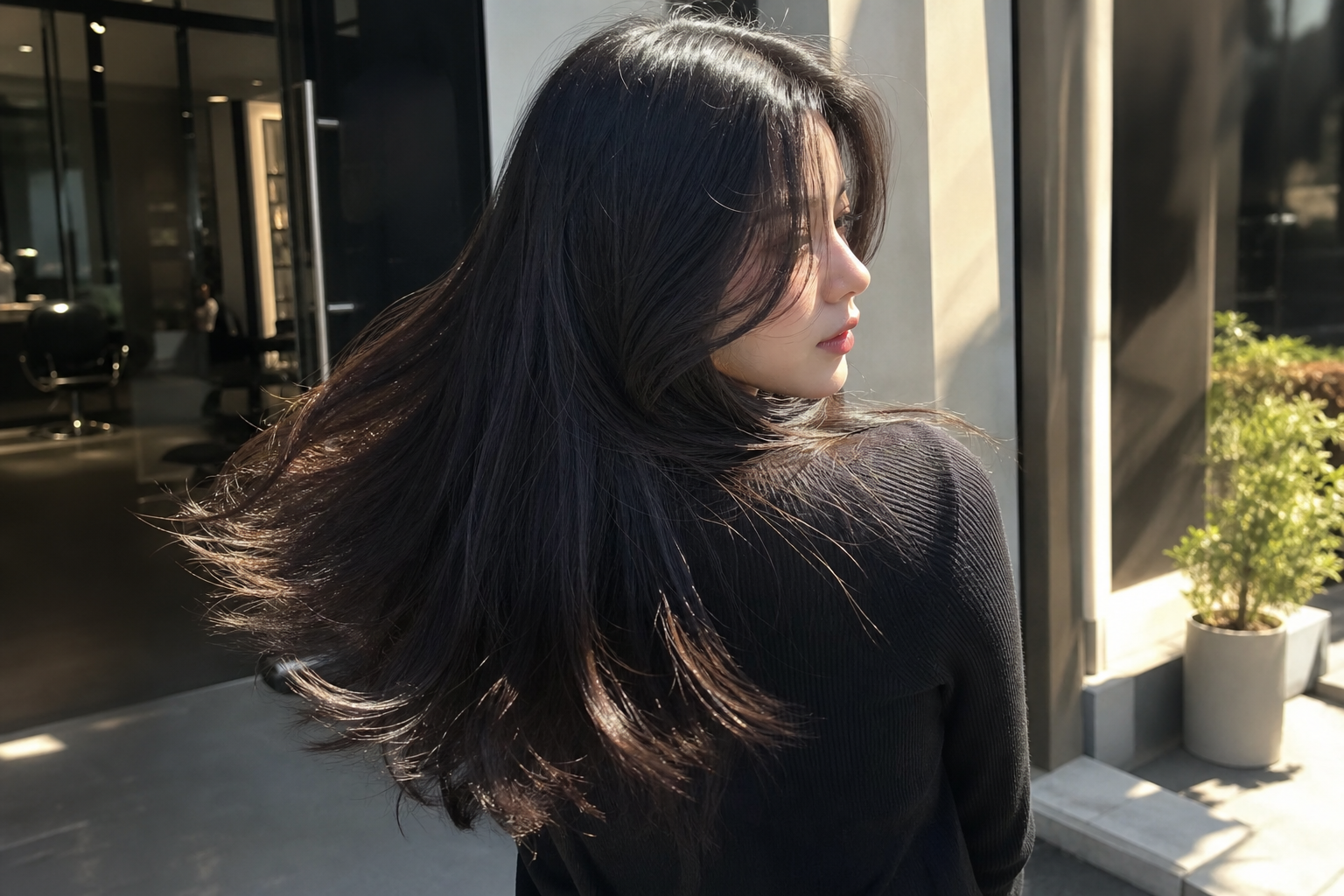 Người phụ nữ đứng ngoài trời với mái tóc đen dài, phần tóc bắt ánh nâu nhẹ dưới nắng