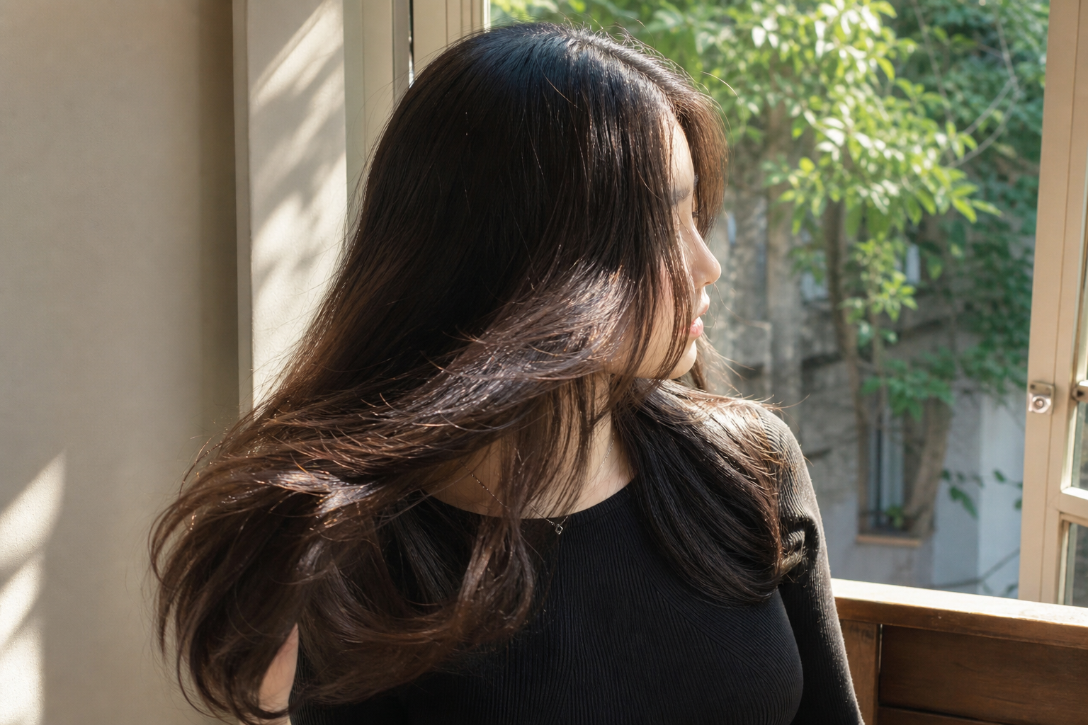 Người phụ nữ tóc nâu đen dài đứng cạnh cửa sổ, tóc bắt sáng nhẹ dưới nắng và nhìn ra ngoài