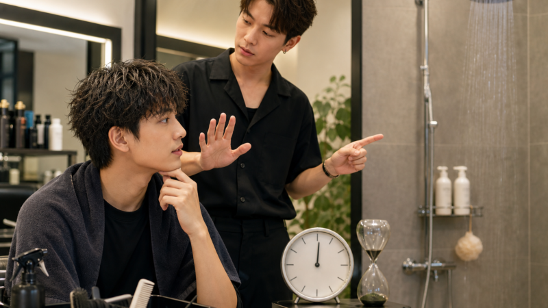 Hai người đàn ông trong salon tóc, một người ngồi trước gương với tóc ướt, người kia đứng cạnh chỉ tay về phía khu vực tắm và đồng hồ cát.