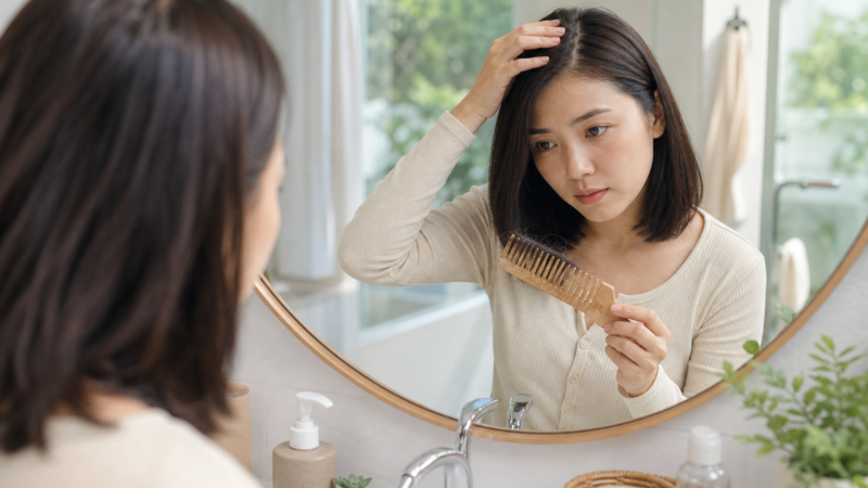 Người phụ nữ soi gương, tay chạm tóc và cầm lược gỗ trong phòng tắm sáng, trông như đang kiểm tra tóc rụng sau sinh tại nhà.