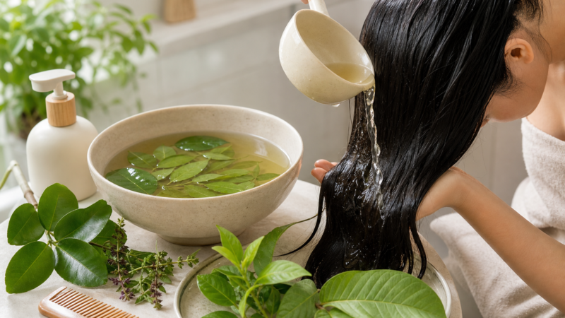 Người phụ nữ đang dội nước lá lên mái tóc dài đen ướt, bên cạnh bát nước ngâm lá và một chiếc lược trên bàn.